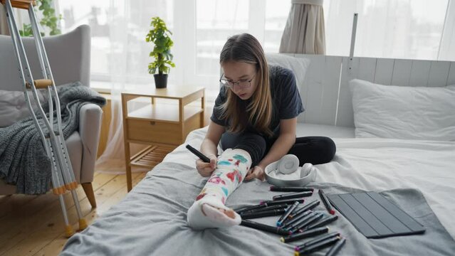 Teenage girl after injury with broken leg in orthopedic cast painting pictures felt tips sitting in bed at home. Teen in glasses drawing on cast. Recovery of damaged leg, forced sick leave concept.
