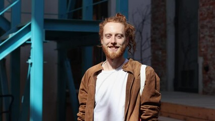 Young man with long, curly red hair and a beard free his hair outdoors. Handsome guy wear rainbow colored accessories.