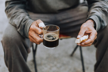 An old, elderly, homeless, poor male worker holds a glass cup of coffee, a croissant, a pastry during lunch. Close up food photography, poverty concept.