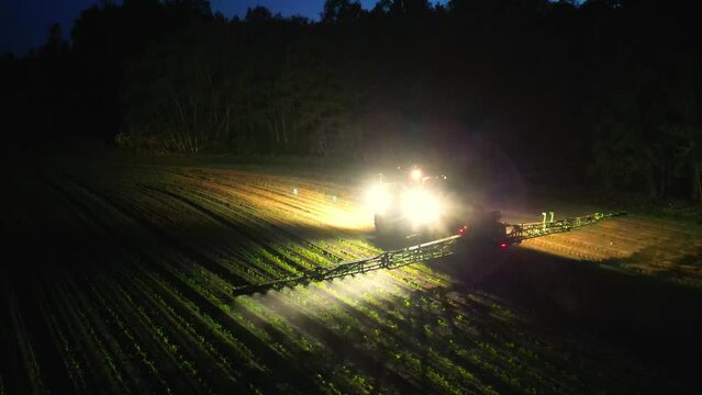 Tracktor in field after sunset. Aerial view of harvesters spraying pesticide. Aerial view of combine harvester working at night. Illuminated field at night during the end of harvesting season