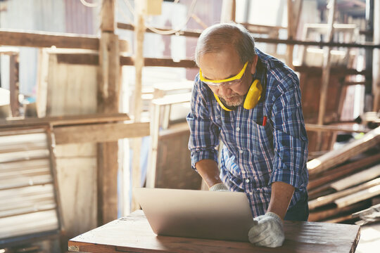 Senior Asian male carpenter working with laptop at furniture workshop. Asian male joiner using laptop computer at woodwork workshop