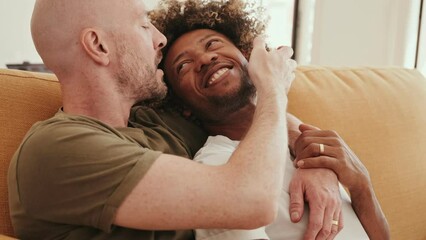 Caucasian and African same-sex couple lovingly embrace on a cozy sofa at home, expressing admiration and joy together - Powered by Adobe