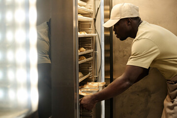 Side view portrait of young male baker putting fresh pastries in freezer at bakery kitchen, copy...