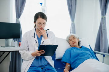 Doctor in white suit take notes while discussing and Asian elderly, woman patient who lying on bed with receiving saline solution in hospital or clinic.