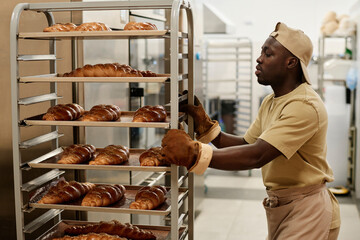Side view portrait of Black young man moving shelves with freshly baked breads in bakery kitchen, copy space