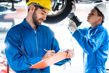 Caucasian automotive mechanic man writing on maintenance document clipboard and checking under car in auto repair garage, assistant pointing flash light, transport business and after service concept