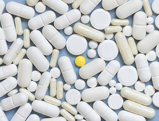 Mix of white medical capsules and pills with one yellow pill in the middle on light blue top view