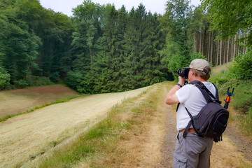 Rear view of senior adult male photographer taking photo of green leafy trees on blurred background, Wallfharte Weidingen hiking trail, open land for agriculture, sunny spring day in Utscheid, Germany