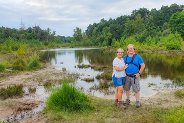 Senior adult couple of hikers standing at muddy bank of a stream among wild vegetation and leafy...