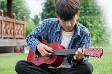 Obraz premium Asian cute boy playing guitar in the front yard of his own house happily, soft focus, concept for recreational activity and free times hobbies of children around the world.