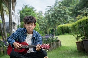 Asian cute boy playing guitar in the front yard of his own house happily, soft focus, concept for recreational activity and free times hobbies of children around the world.