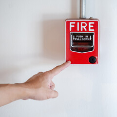 Emergency fire warning system pull station. Hand pointing at red fire alarm mounted on white wall...
