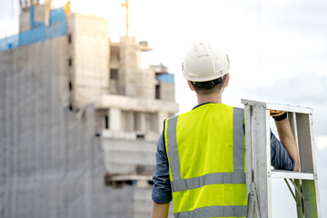 Asian man construction worker with green reflective vest and white safety helmet carrying aluminium ladder looking at unfinished high-rise building at construction site