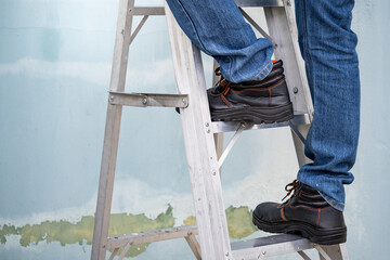 Male maintenance worker feet with protective safety shoes climbing up on aluminum step ladder at...