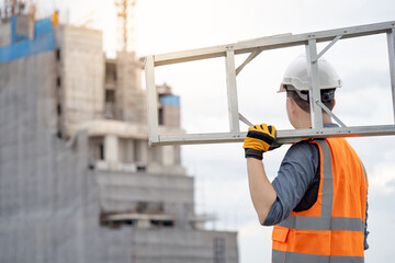 Asian man construction worker with orange reflective vest and white safety helmet carrying aluminium ladder looking at unfinished high-rise building at construction site © Summer Paradive