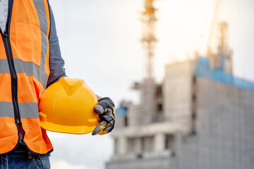 Safety workwear concept. Male hand holding yellow safety helmet or hard hat. Construction worker man with reflective orange vest and protective gloves standing at unfinished building with tower crane