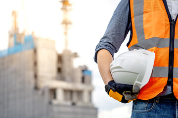 Safety workwear concept. Male hand holding white safety helmet or hard hat. Construction worker man with reflective orange vest and protective gloves standing at unfinished building with tower crane - obrazy, fototapety, plakaty