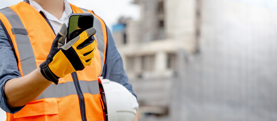 Construction worker man with orange reflective vest holding white protective safety helmet using smartphone at unfinished building site. Male engineer or foreman stay connected with social media app - obrazy, fototapety, plakaty