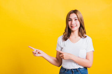 Asian Thai happy portrait beautiful cute young woman standing wear white t-shirt pointing finger away side looking to camera, studio shot isolated on yellow background with copy space