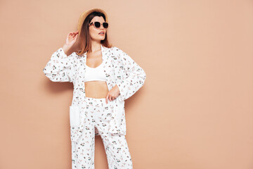 Young beautiful smiling female in trendy summer costume clothes. Carefree woman posing near beige wall in studio. Positive model having fun indoors. Cheerful and happy. In hat and sunglasses