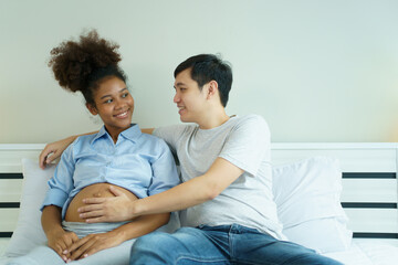 Happy cheerful husband and pregnant wife staying - relaxing together in bedroom, husband embracing or playing on pregnant wife tummy. Diverse ethnicity husband and wife lifestyles concept.