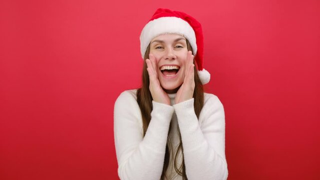 Happy young woman wear white cozy sweater and Santa Claus hat scream sharing hot news about sales, hands near mouth, posing isolated over pastel red studio background. New Year 2024 holiday concept