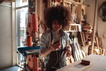 Young mixed woman listening to music on her smartphone in a carpenters garage