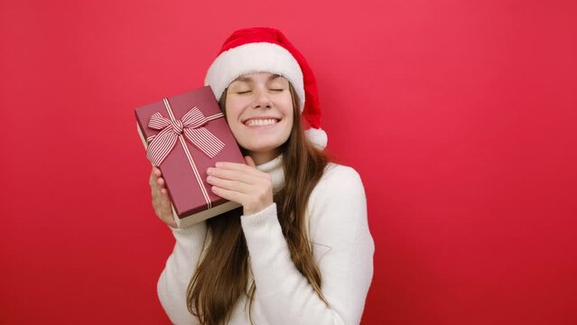 Charming merry young woman wear white sweater and Xmas Santa hat hug present box with gift ribbon, posing isolated over pastel red studio background. Happy New Year 2024 celebration holiday concept