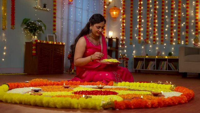 happy young Indian woman preparing diwali flower rangoli on floor for pooja or ritual at home - concept of festive celebration, special occasion and traditional culture or wear