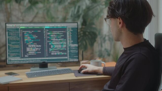 Professional male programmer sitting at desk in home office, looking at PC screen and scrolling through computer code