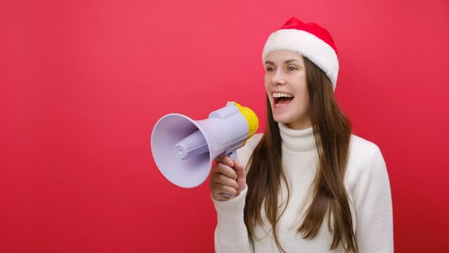 Pretty young woman wear white sweater and Xmas Santa hat scream in megaphone announces sale, posing isolated over pastel red color background in studio. Happy New Year 2024 celebration holiday concept