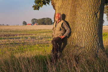An elderly man leans against the mighty trunk of an oak tree, basking in the evening sun, savoring...