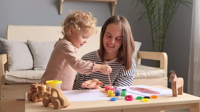 Creative togetherness. Painting love. Maternal inspiration. Childlike wonder. Art bonding. Joyful painting. Overjoyed woman with infant daughter drawing with colorful paints together in living room.