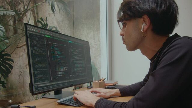 Concentrated software developer in wireless earphones writing computer code on PC at desk in home office