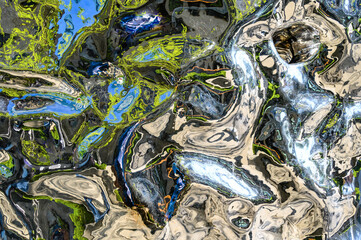Part of a warped, scratched chrome surface with multi-colored objects reflected in it, creating an abstract composition