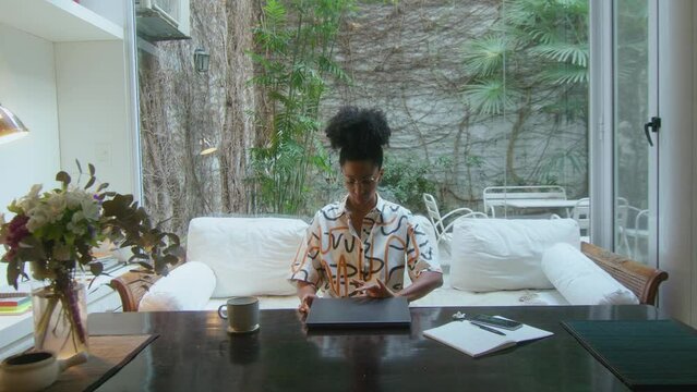 African-American woman walking into modern home office with backyard garden view, sitting on sofa, opening laptop and working online. Front view, zoom shot