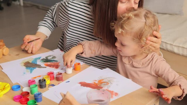 Unknown mother with her little baby daughter painting together in home interior using water color paintbrush and fingers for making creative picture enjoying bonding moments.