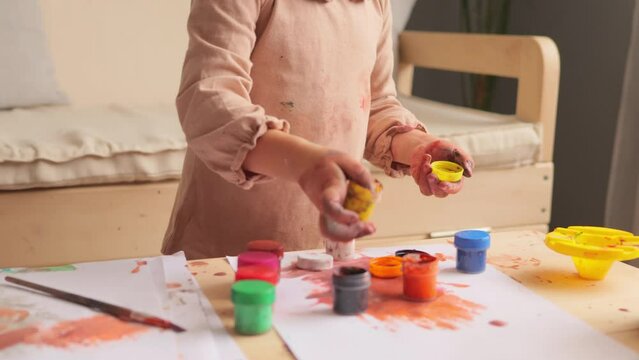 Unrecognizable baby girl learning painting with water colors in light room interior opening paint bottles preparing her workplace for creating amazing pictures.