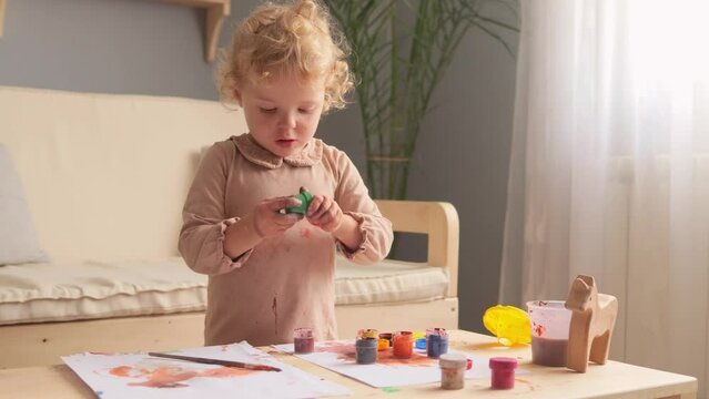 Colorful preschooler learning. Cute toddler's colorful art. Child's education. Blonde curly haired diligent infant child drawing using colorful paints at home or nursery.