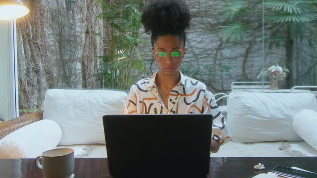 Young black woman sitting in modern home office with backyard garden view, typing laptop and working on a freelance project. Front view, dolly zoom shot