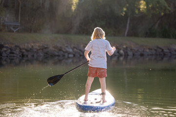 Pre teen boy stand up paddle boarding on lake in golden afternoon light