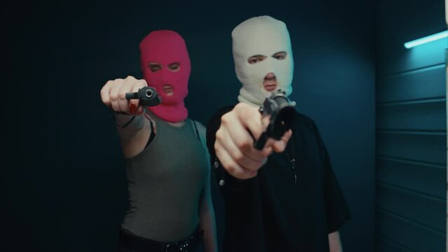 Medium POV shot of young caucasian couple in balaclavas pointing with handguns at camera indoors