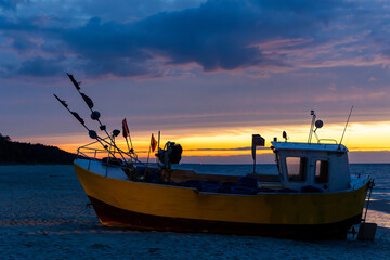 A yellow-orange fishing boat pulled up on the beach during sunset. Dramatic sunset sky with clouds. Photo taken in the evening with minimal lighting