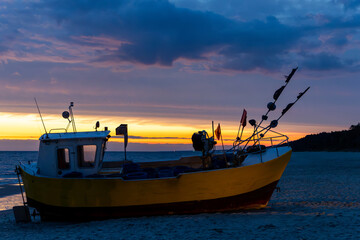 A yellow-orange fishing boat pulled up on the beach during sunset. Dramatic sunset sky with clouds. Photo taken in the evening with minimal lighting