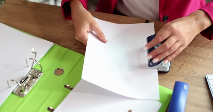 Female hands of accountant or secretary are punching holes with hole punch in documents in office. Concept of stationery and business archive