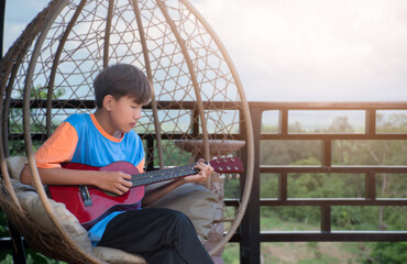 Asian cute boy sitting and playing the guitar on the swing bird nest in the backyard of his house, recreational  activity, free times activity and hobby of boys around the world concept.