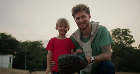 Portrait of a Happy little blond boy with blue eyes in a red T-shirt who stands with his dad with curly hair with stubble in a Green T-shirt and wearing a baseball glove in the park - Powered by Adobe