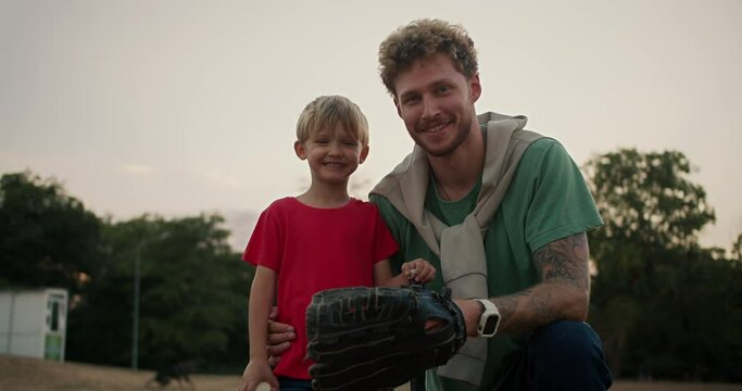 Portrait of a Happy little blond boy with blue eyes in a red T-shirt who stands with his dad with curly hair with stubble in a Green T-shirt and wearing a baseball glove in the park