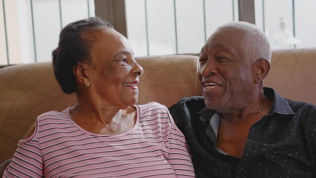 A Happy Black Senior Couple Sitting On Couch Looking At Each Other Conversing. African American Husband And Wife In Old Age Relationship