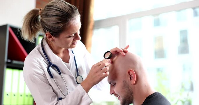 Trichologist with magnifying glass examines bald patient with alopecia in clinic. Hair growth concept of hair loss in man
